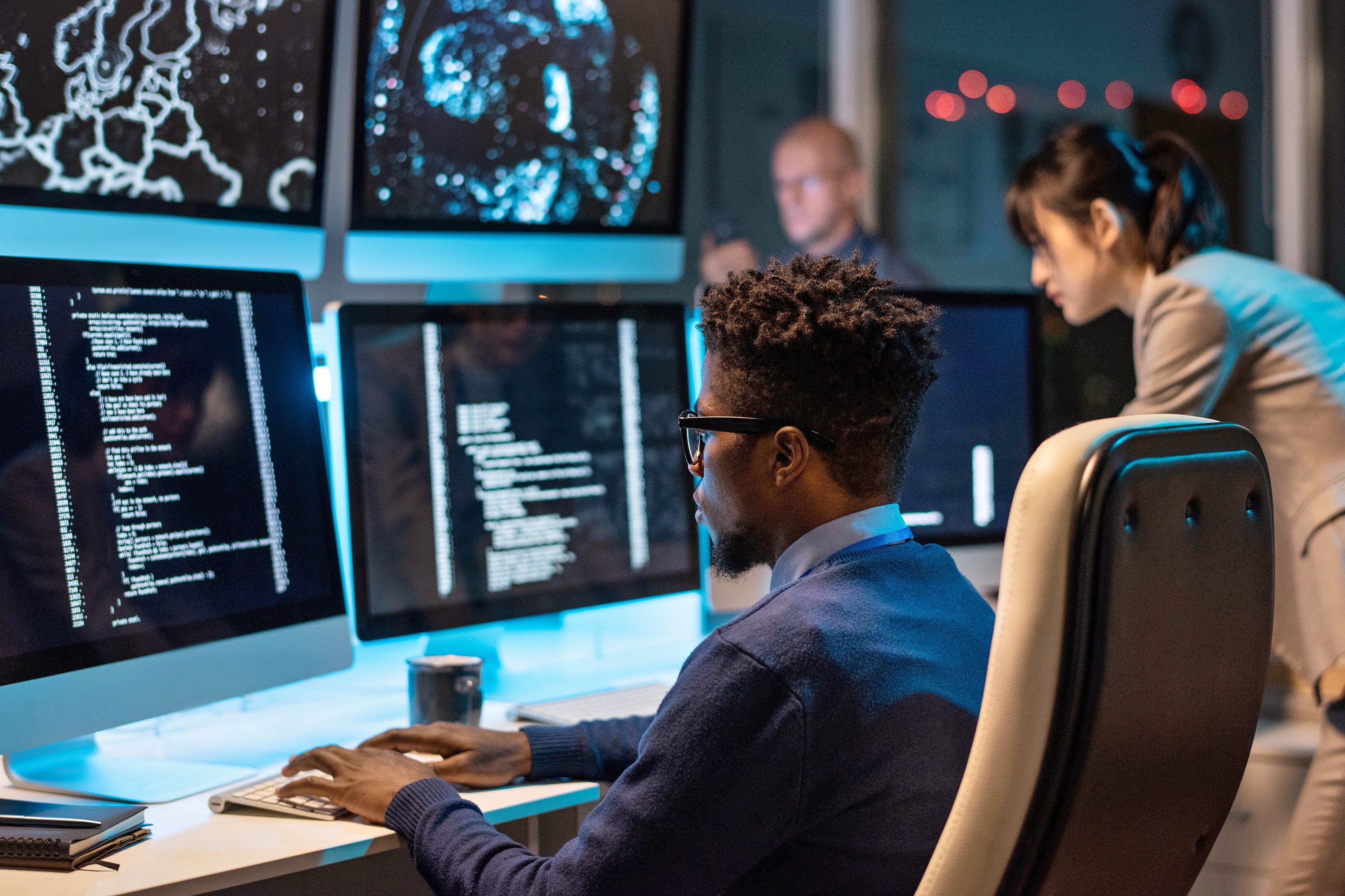 A security analyst works diligently at his desk, surrounded by multiple monitors displaying lines of code and network diagrams, representing the thorough data security assessment services provided by Fast Fixx.