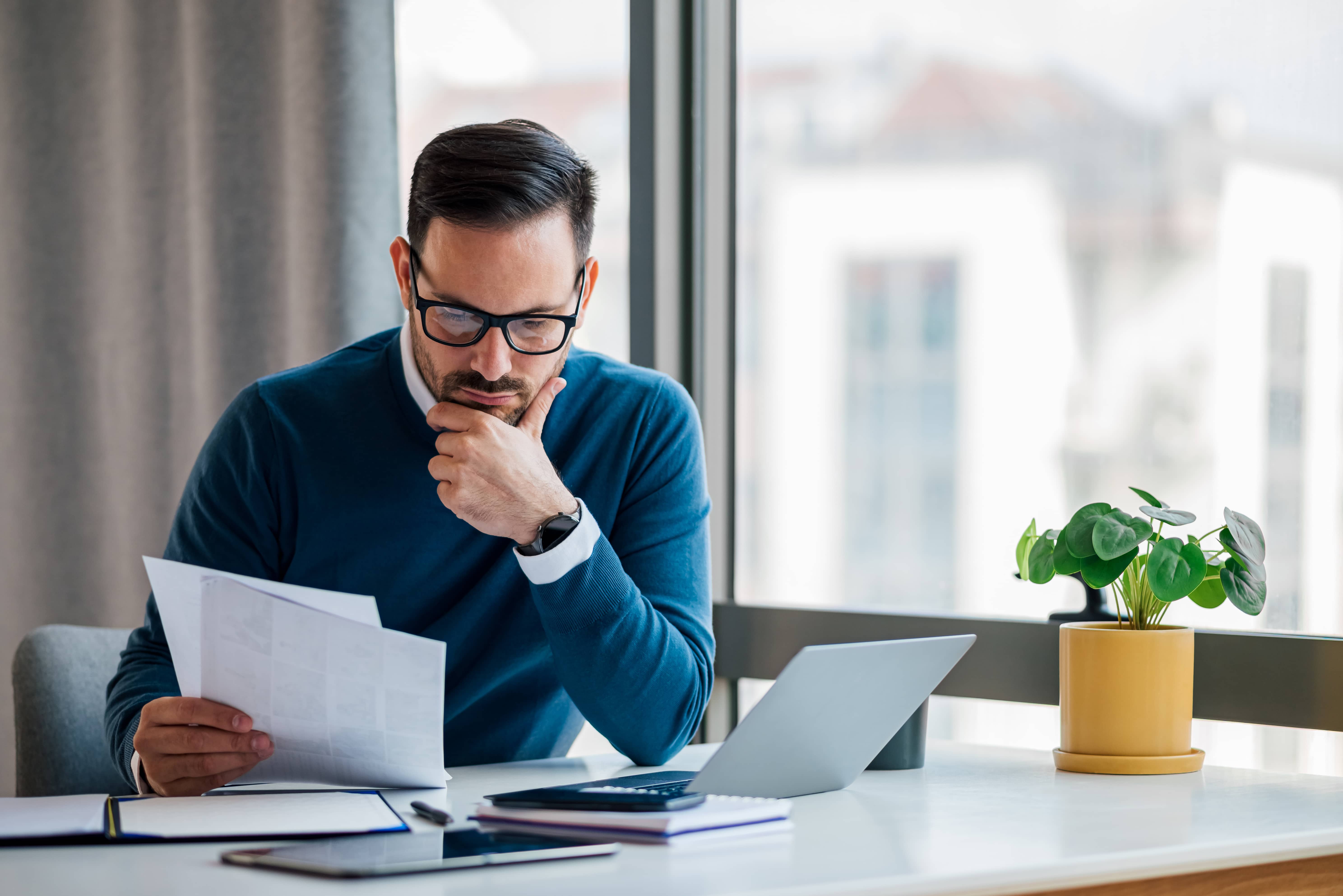 A man in glasses sits at his desk, reviewing a document while in deep thought, symbolizing the development and implementation of a customized compliance program tailored to a specific industry and its needs.