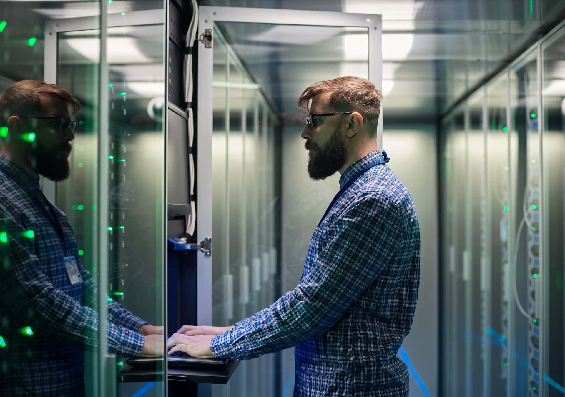 A bearded IT professional in a server room, working on a laptop to monitor network activity and detect potential intrusions