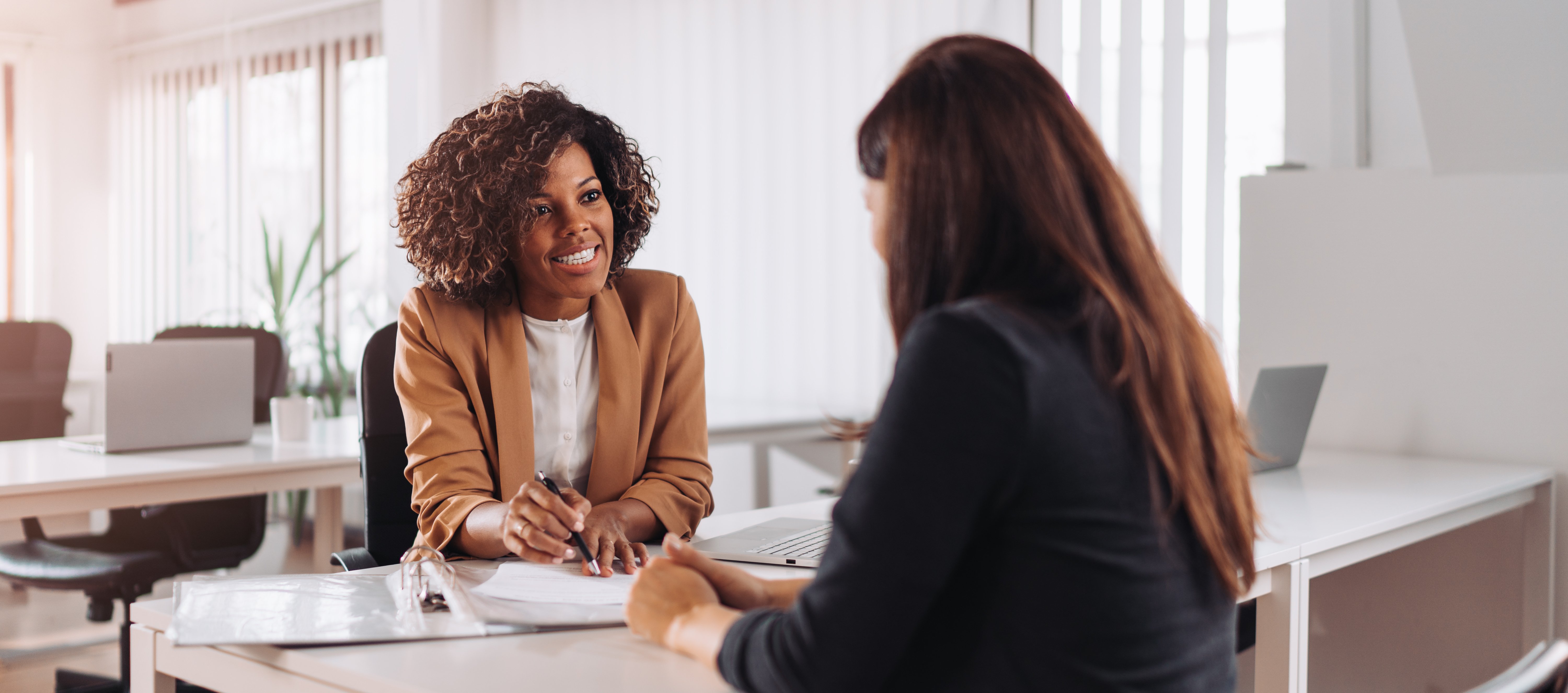 A smiling woman in a brown blazer is having a meeting with a client, discussing compliance consulting and auditing services to achieve and maintain regulatory and data privacy standards.